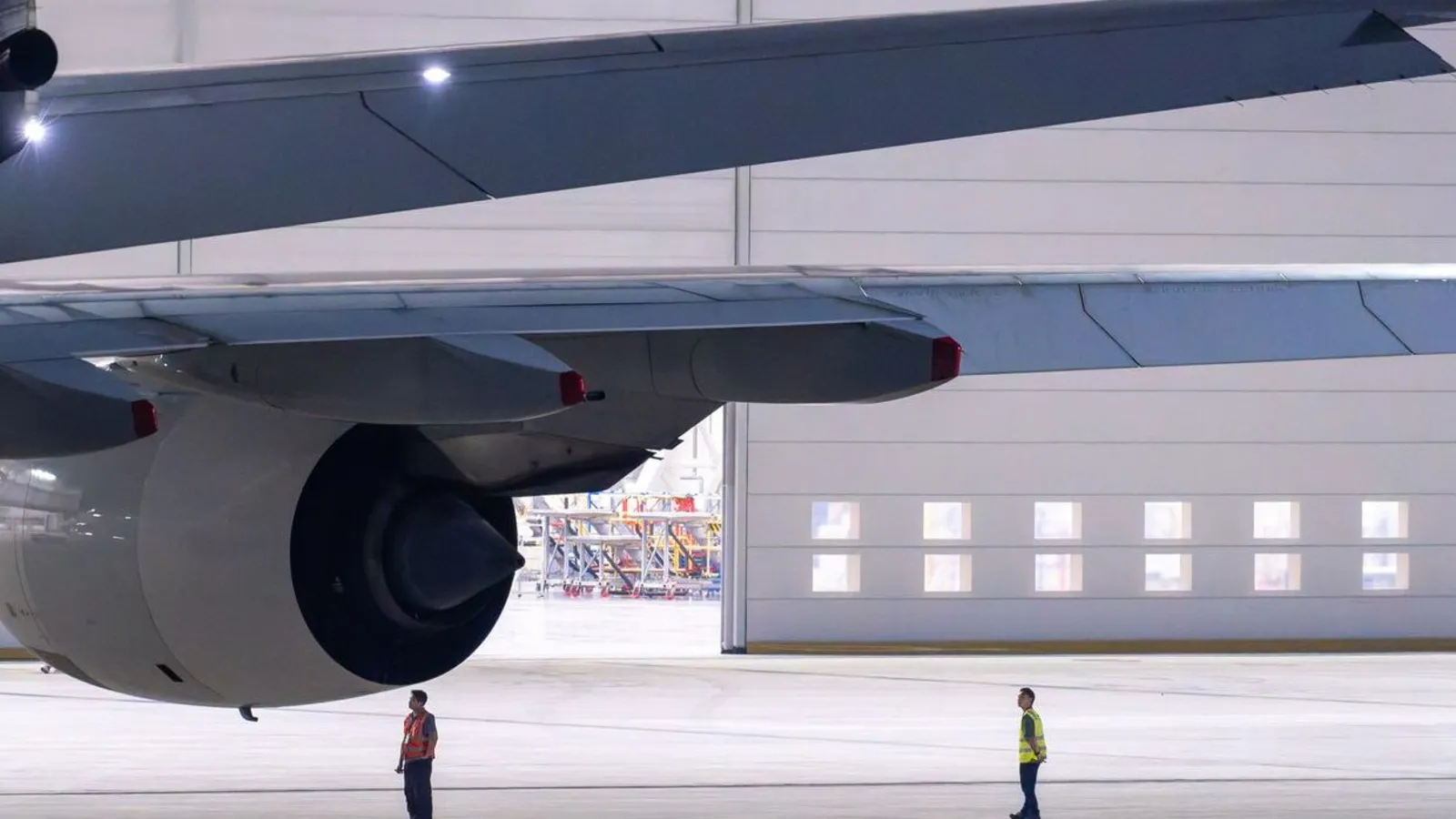 Two ground crew members in high-visibility vests stand under the wing and engine of a jet inside a large maintenance hangar